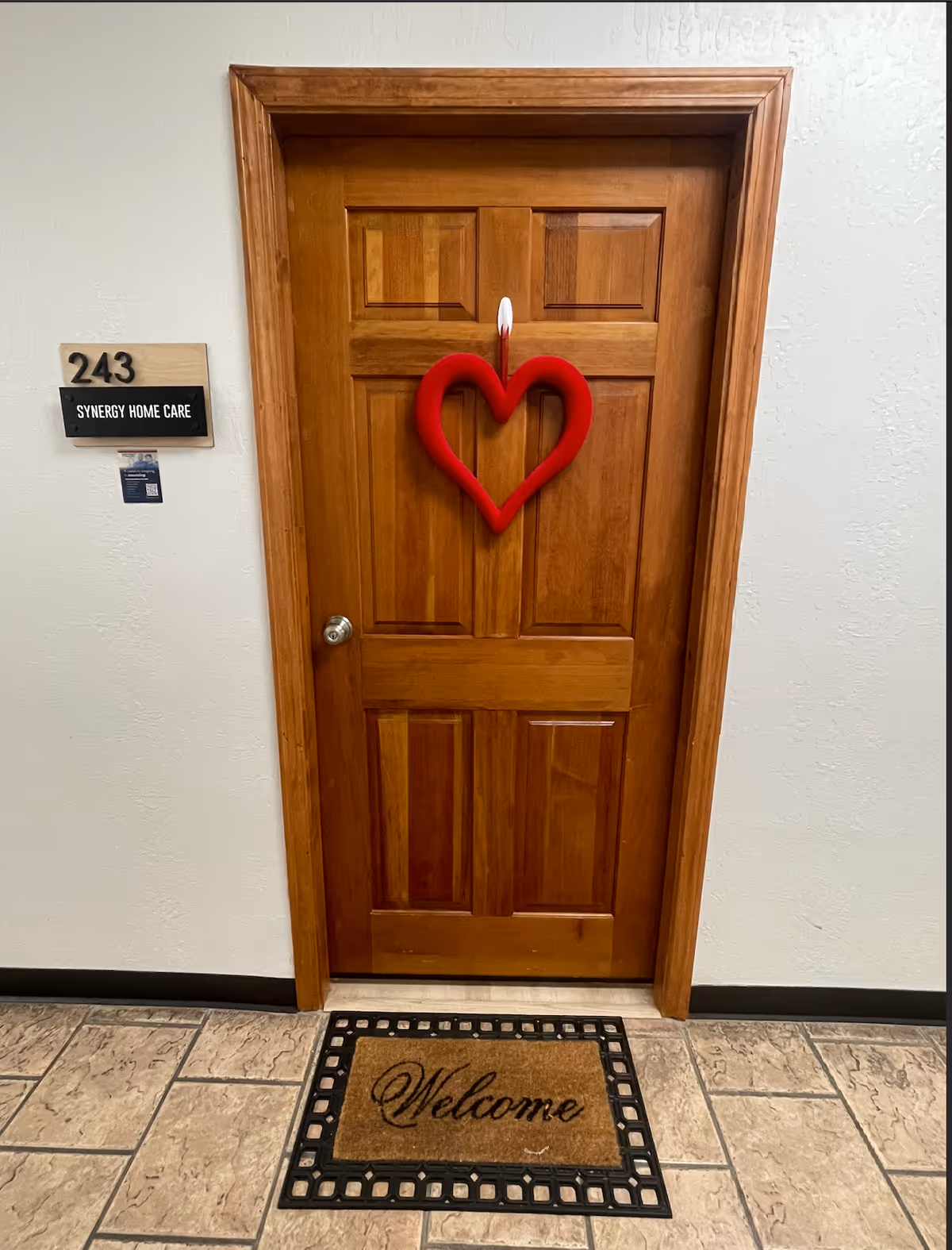 Wooden door with a red heart wreath in a hallway, signed Synergy Home Care, welcome mat.