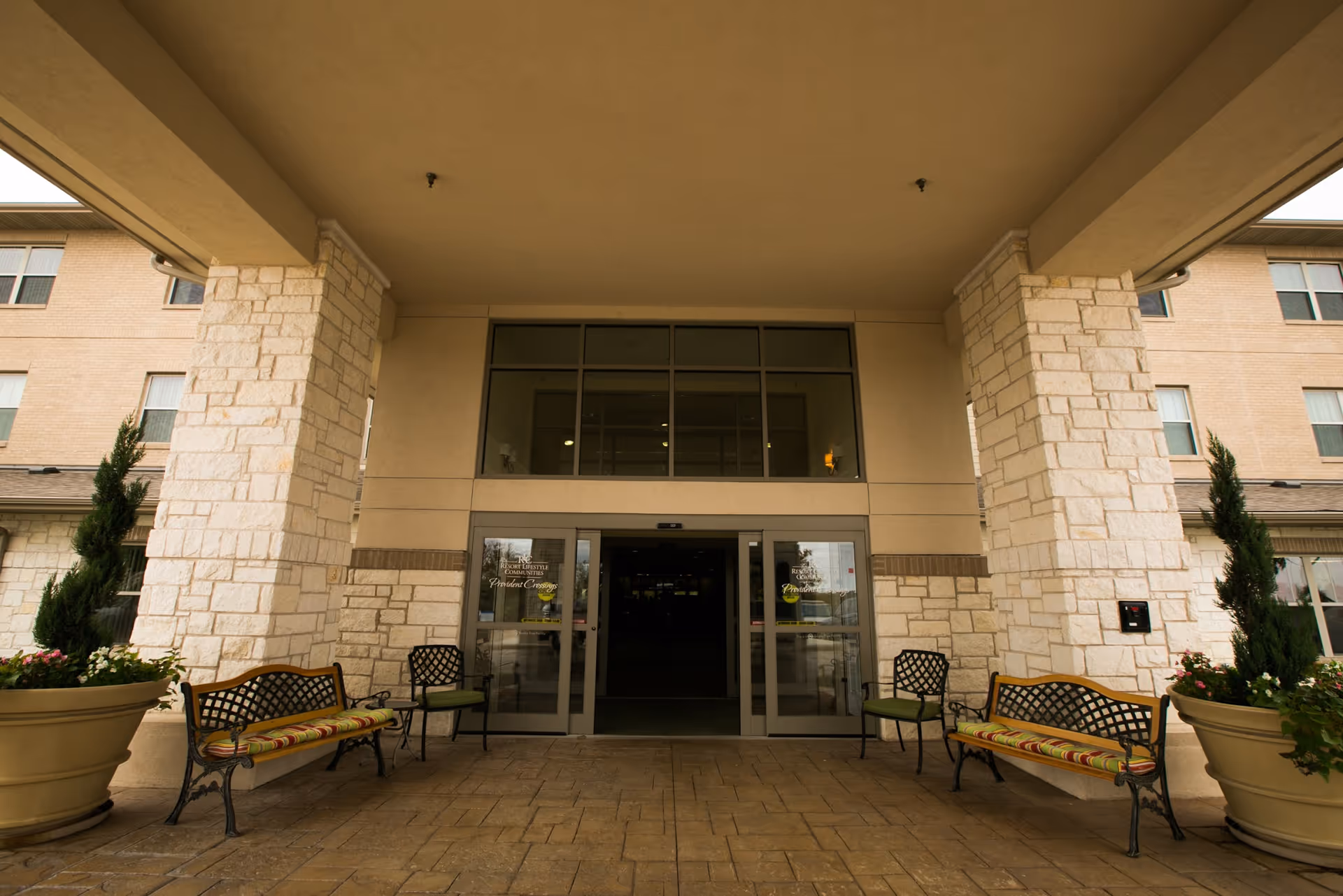 Entrance of Provident Crossings Retirement Resort showing a covered driveway with stone pillars, two large planters with greenery, and benches with cushions on either side of the automatic glass doors.
