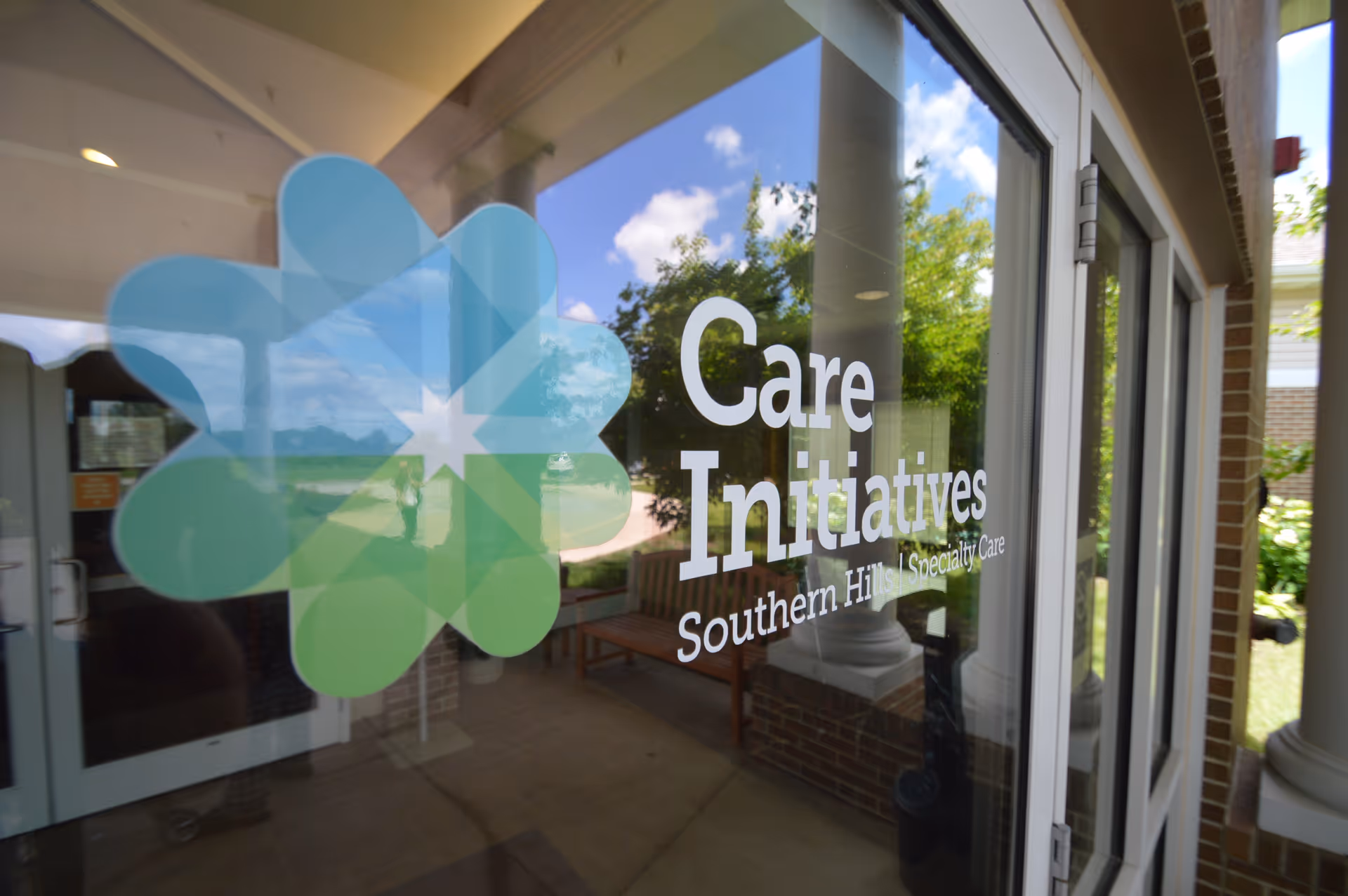 Glass door with a colorful flower-shaped logo and the text 'Care Initiatives Southern Hills Specialty Care' printed on it, with a reflection of the outdoor area including trees, columns, and a bench.