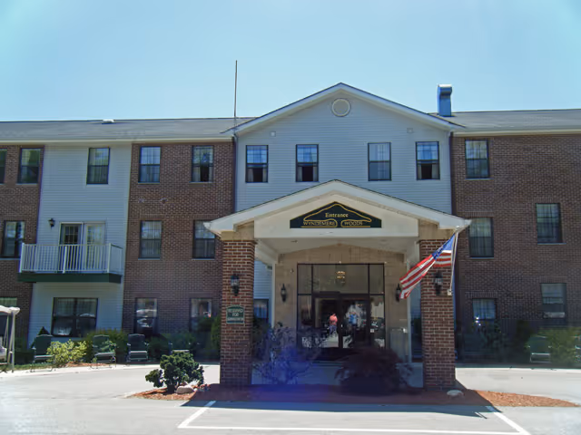 Front exterior view of a three-story senior living facility named Wyndemere Woods with a covered entrance, brick and siding facade, and an American flag displayed near the entrance.