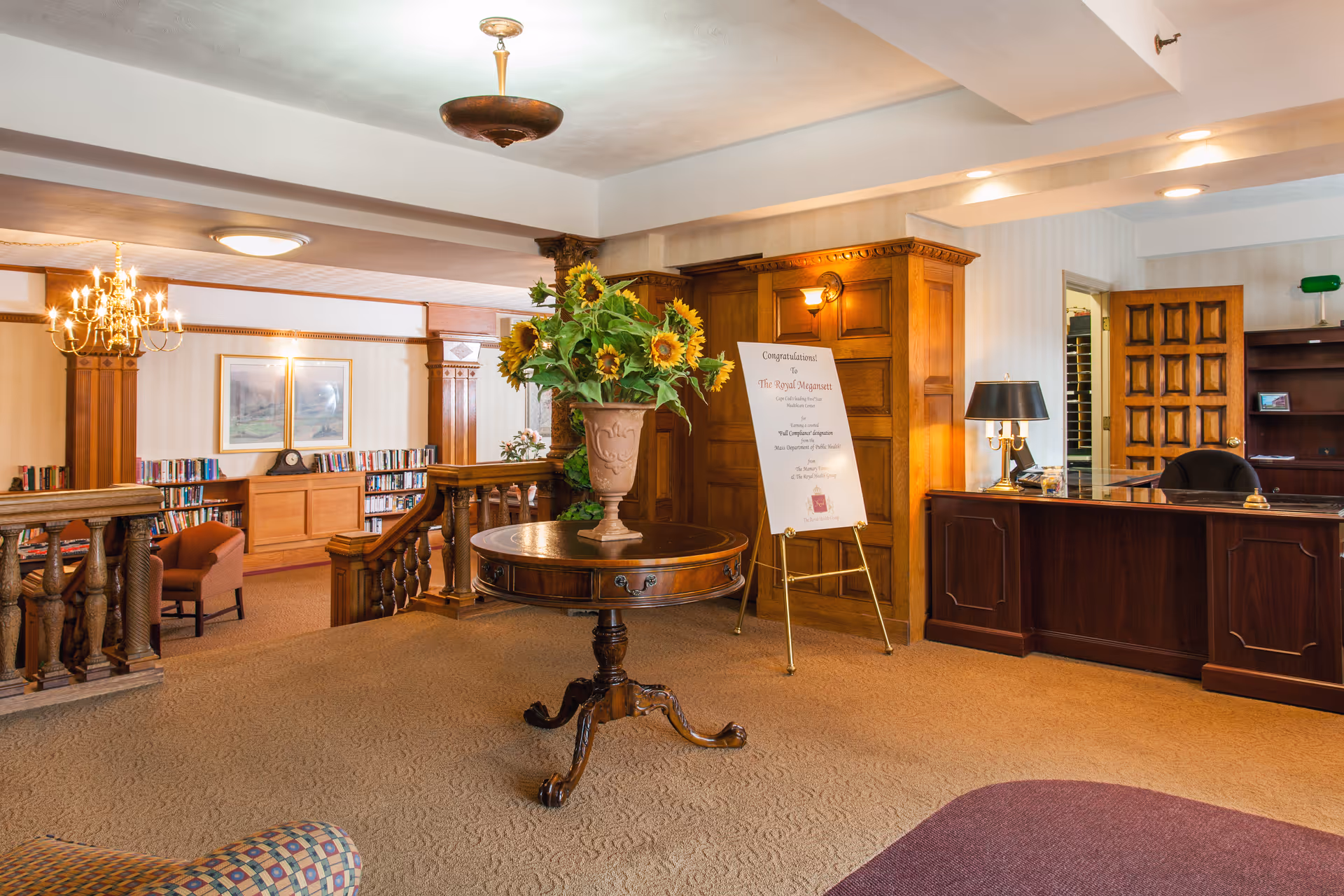 A warm nursing home lobby with a round table displaying a vase of sunflowers, a wooden reception desk, bookshelves and seating areas.