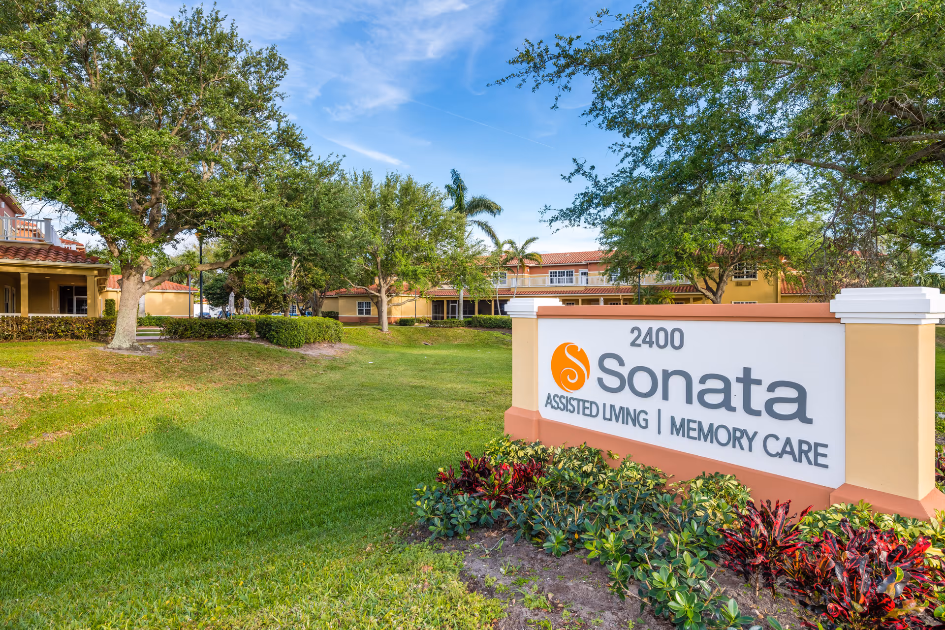 Outdoor view of Sonata Boynton Beach assisted living and memory care facility with a large sign displaying the name and address 2400 Sonata. The area is landscaped with green grass, bushes, and trees under a clear blue sky.