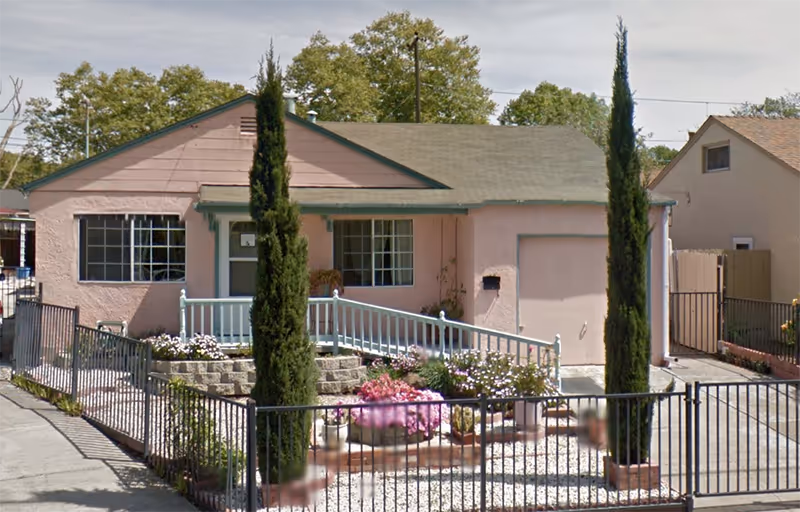 Small pink single-story building with a front garden, fence, and two tall trees.