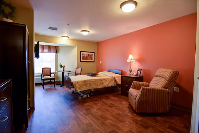 A warm senior living bedroom with a single adjustable bed, armchair, nightstand with lamp, small table and chairs, and wood flooring.