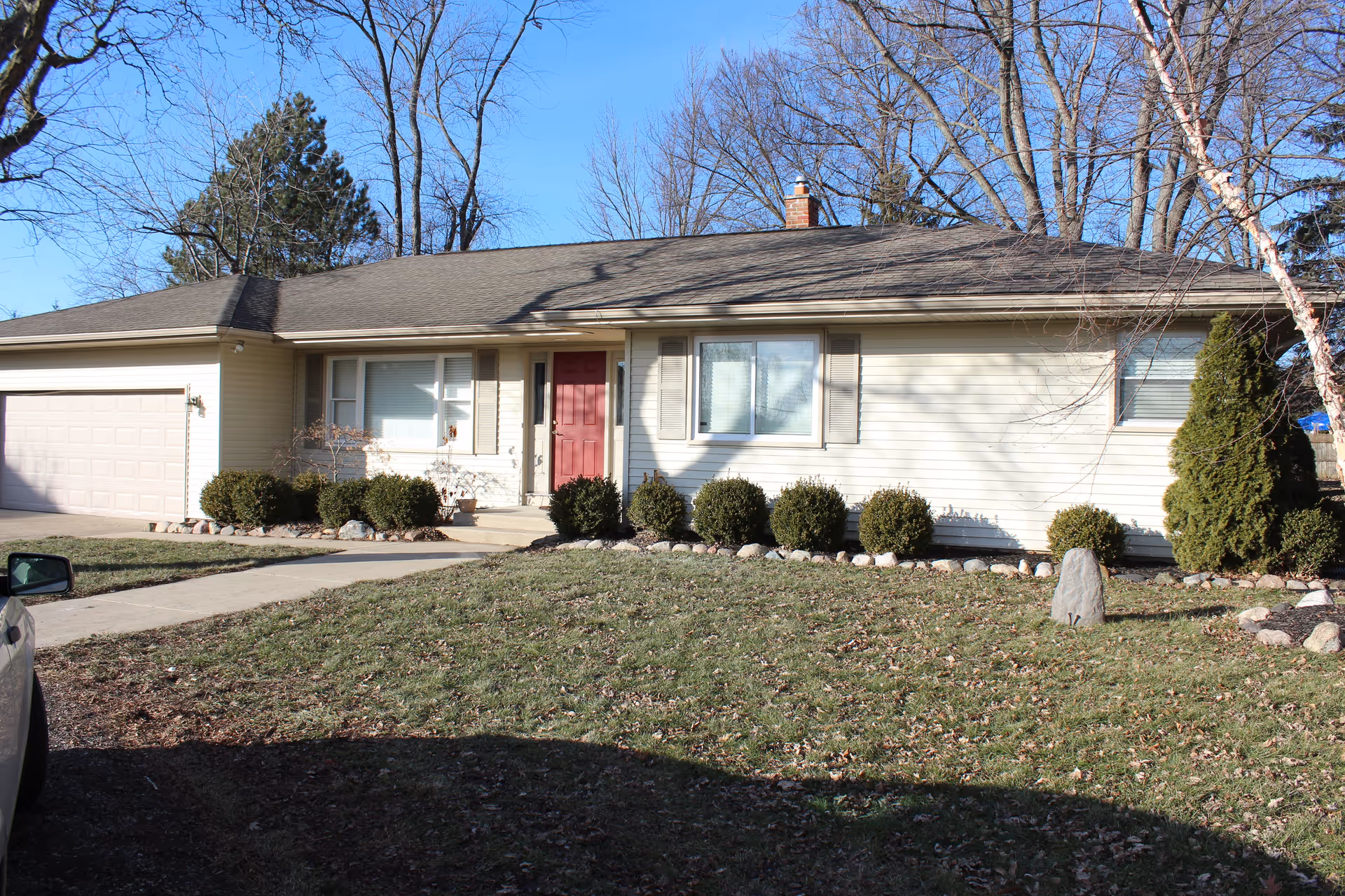 Single-story ranch-style house with a red front door, attached garage, and a grassy front yard with shrubs.
