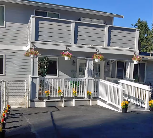Exterior view of a gray senior care facility with a white porch and hanging baskets.