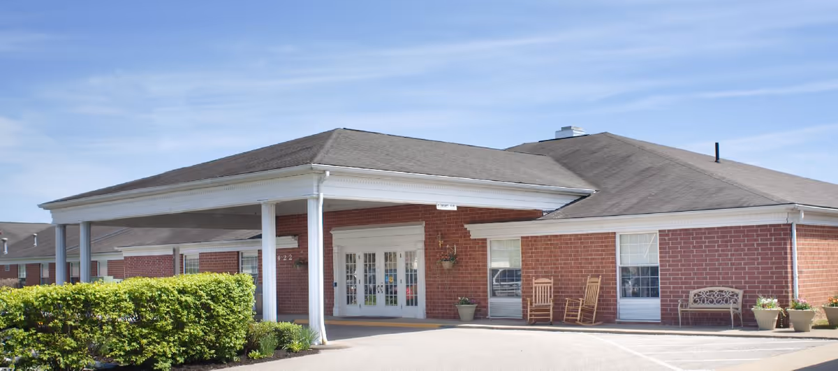 Exterior view of a single-story brick building with a covered entrance supported by white columns. There are two rocking chairs and a bench near the entrance, along with some potted plants. The sky is clear and blue.