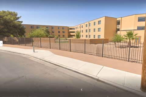 Exterior view of a beige multi-story senior living facility with a metal fence along a sidewalk and desert landscaping.