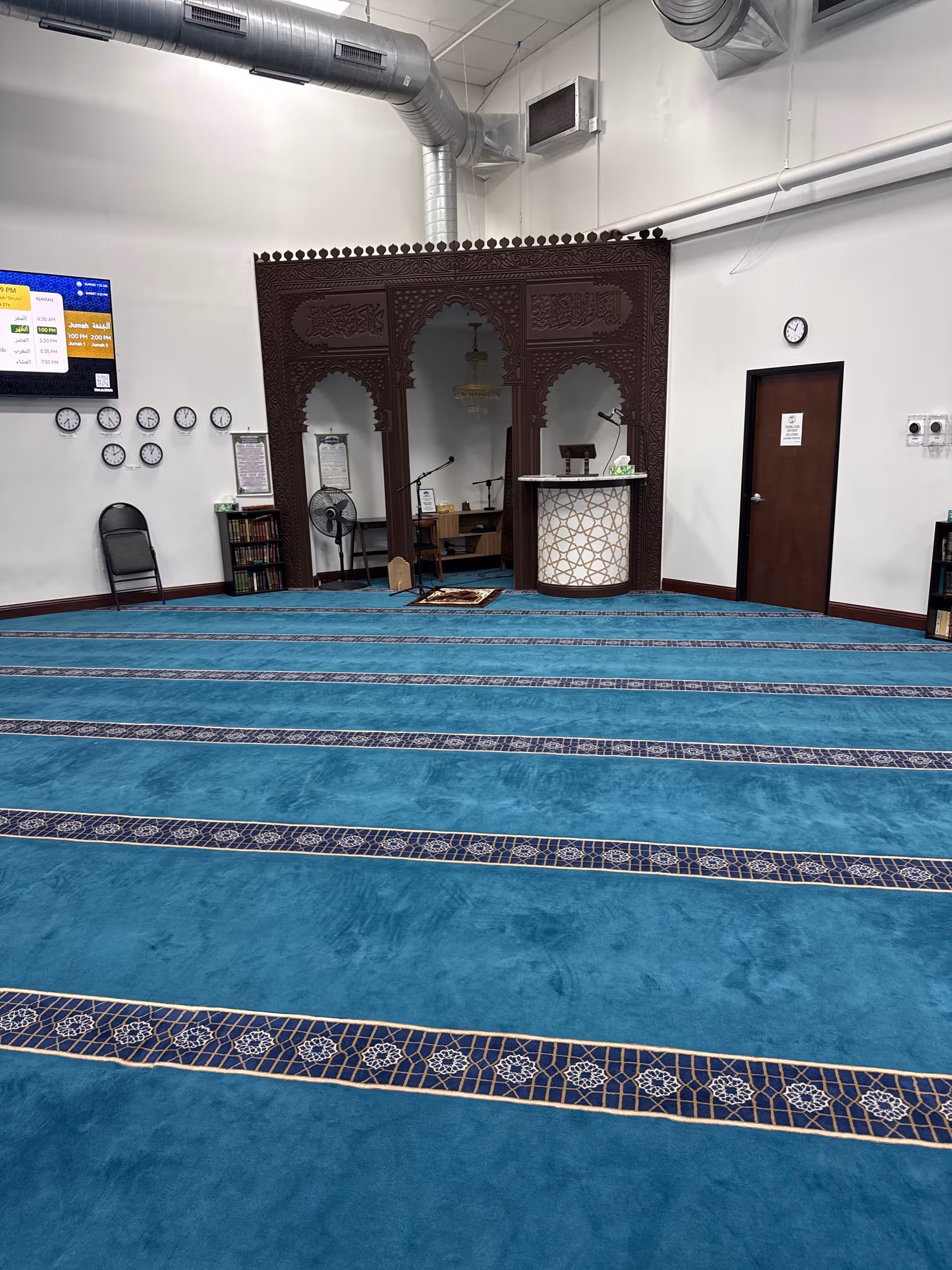 Blue carpeted prayer room with ornate wooden arch divider and podium inside Xmed facility.