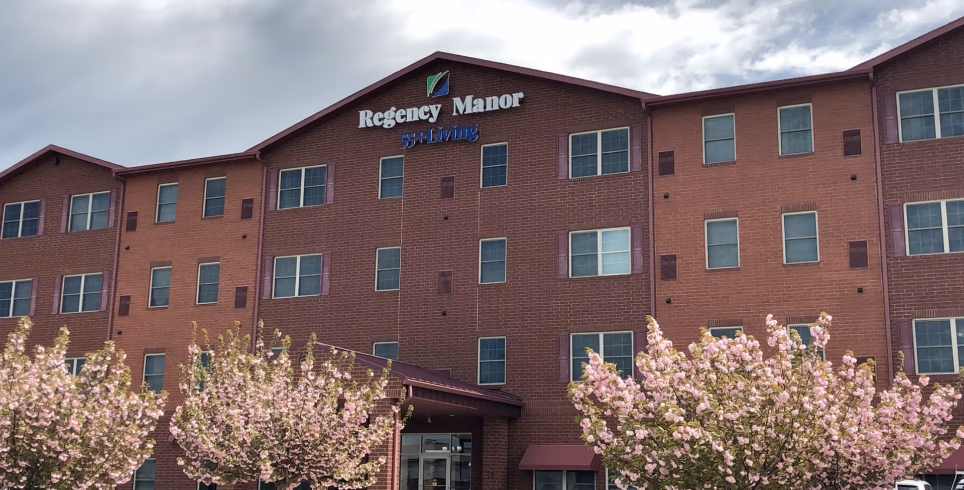 Exterior view of a multi-story brick building with multiple windows and a sign that reads 'Regency Manor 55+ Living'. In front of the building, there are blooming pink flowering trees under a partly cloudy sky.