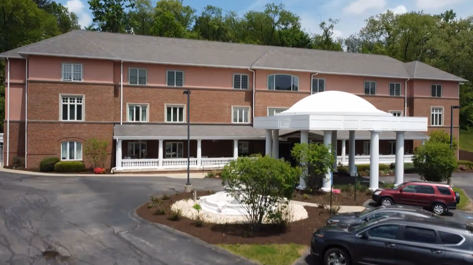 Exterior view of a three-story senior living facility building with a brick and pink facade, a large white covered entrance supported by columns, surrounded by trees and parked cars in the driveway.
