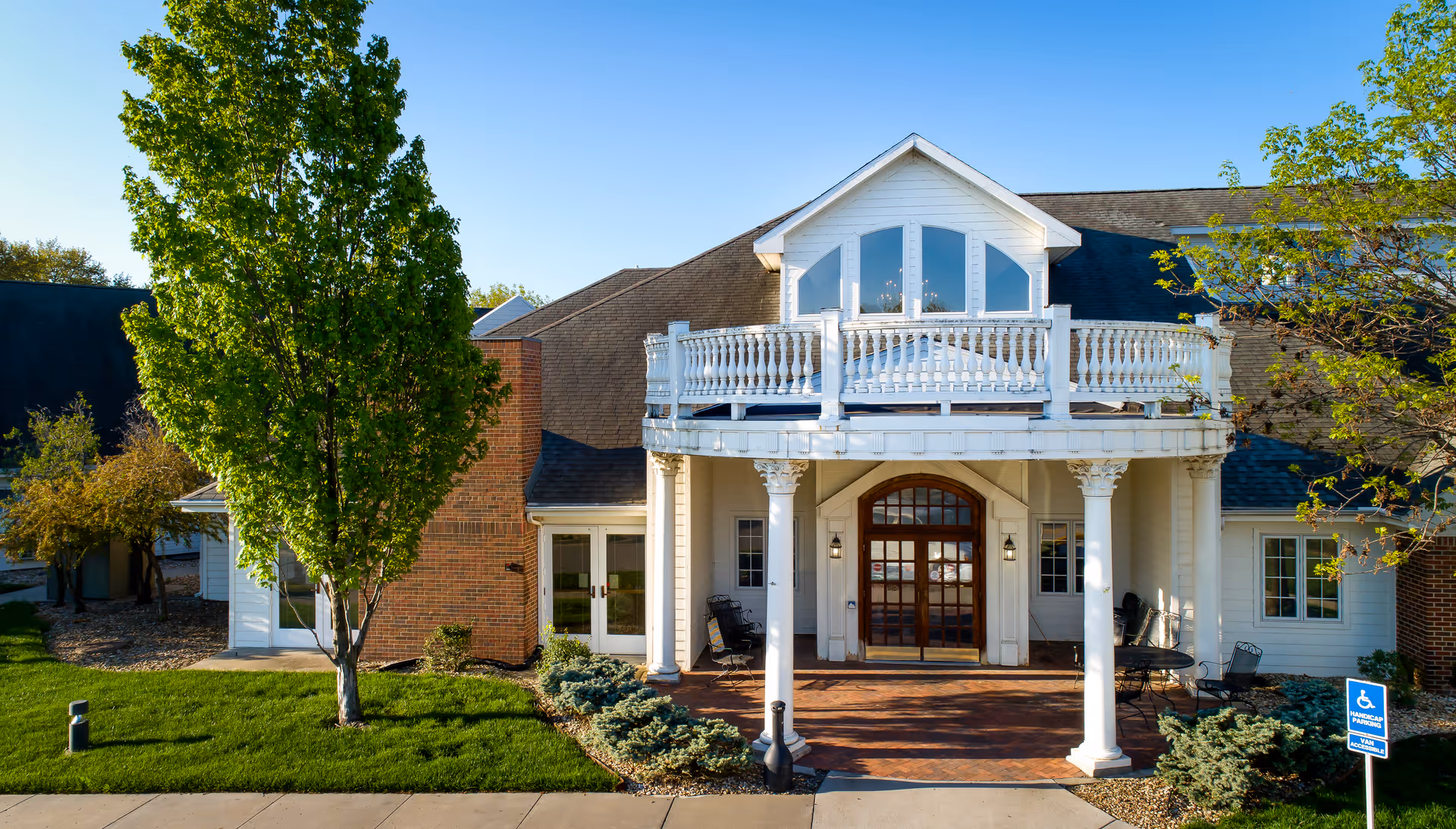 Front exterior view of Lexington Park Health & Rehab of Topeka showing a building entrance with white columns, a balcony with white railing, large windows, and a brick pathway. There are trees and shrubs on either side of the entrance and a blue handicap parking sign visible on the right.