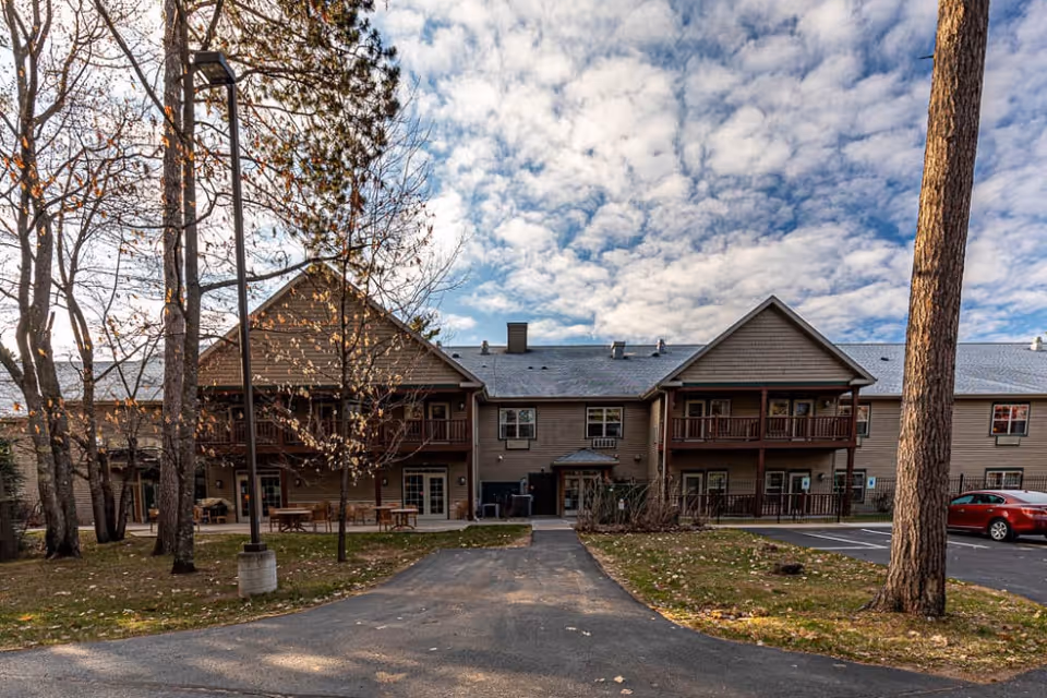 Exterior view of a two-story senior living facility building with balconies and a parking area. The building is surrounded by trees with some fallen leaves on the ground under a partly cloudy sky.