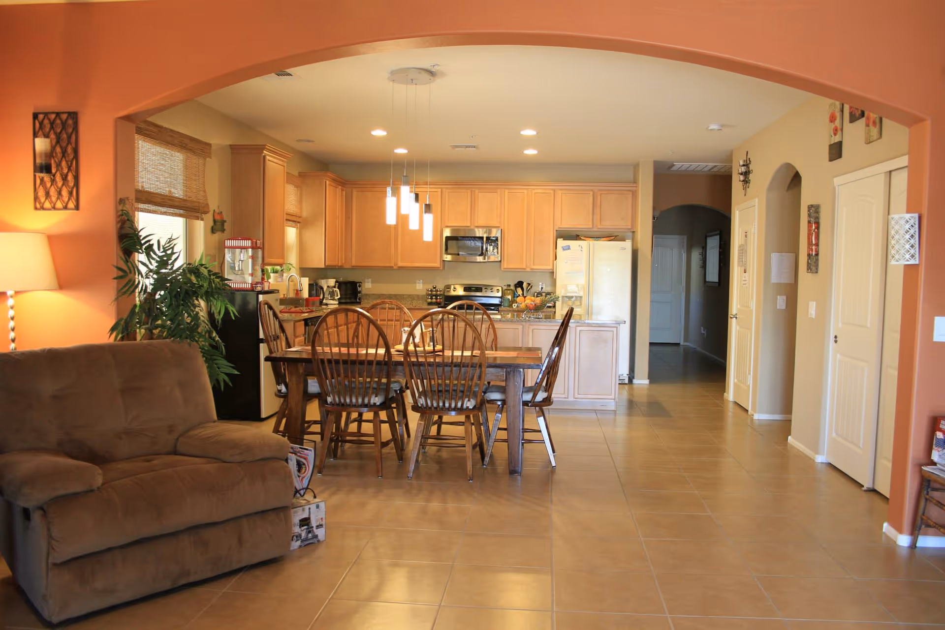 Open-concept living area showing a wooden dining table with chairs in front of a kitchen with light wood cabinets and tiled floors.