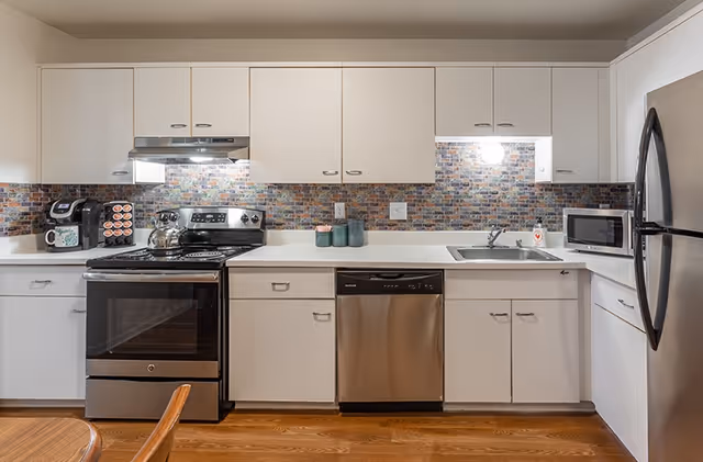 White modern kitchen with stainless steel appliances, mosaic backsplash, and wooden floor.