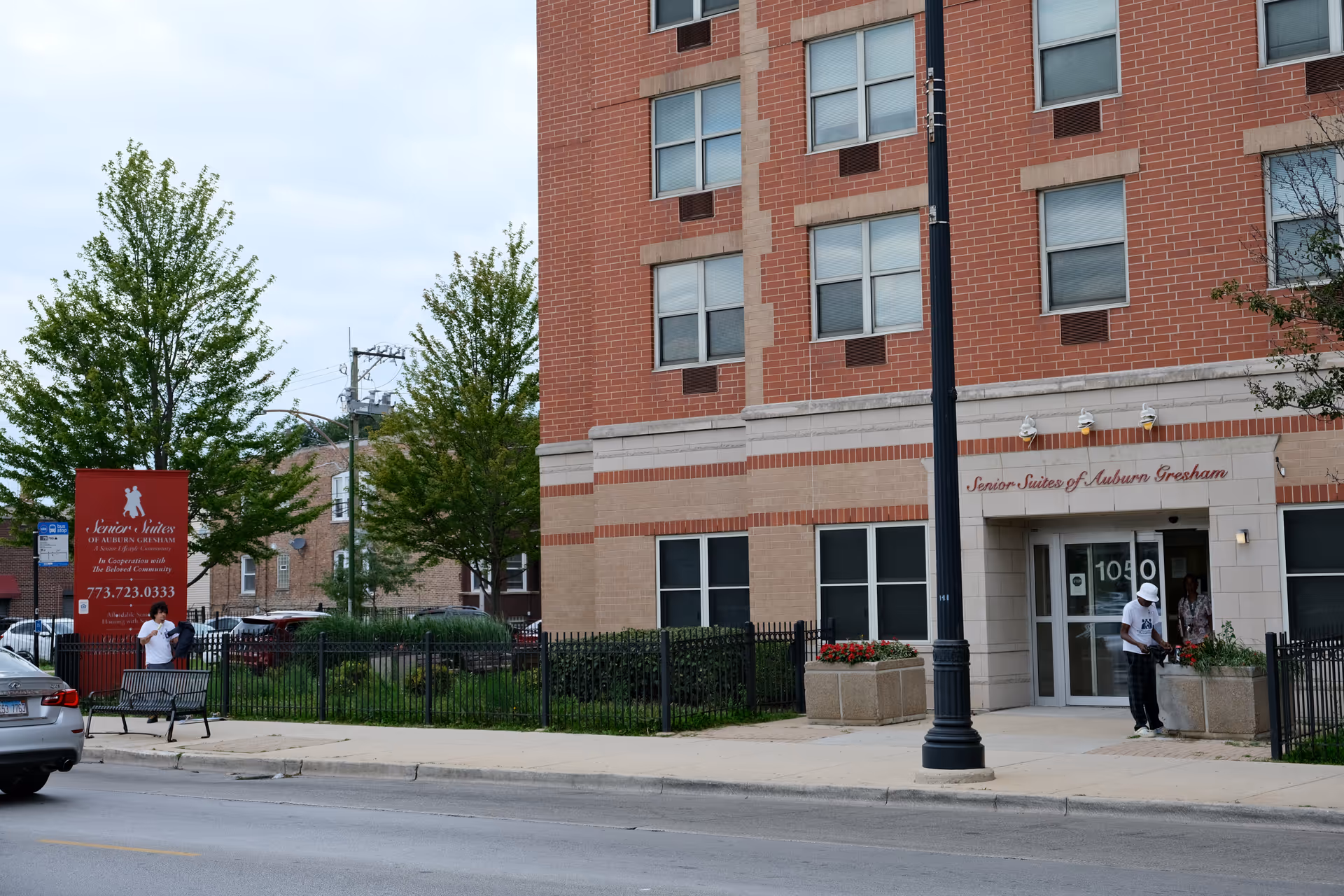 Front entrance of the brick Senior Suites of Auburn Gresham building with a sidewalk, sign, and a few people near the doorway.