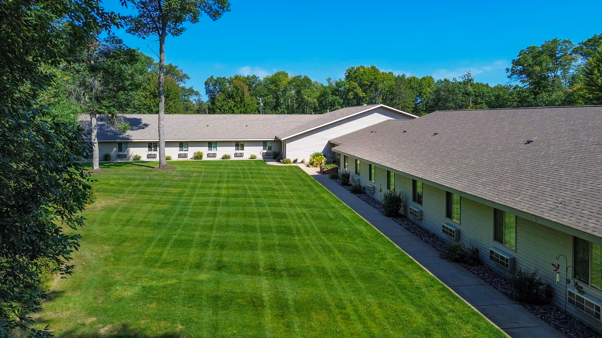 Aerial view of a senior living facility named Golden Horizons of Crosslake, showing two long single-story buildings forming an L shape around a large, well-maintained grassy courtyard with trees and shrubs, under a clear blue sky.