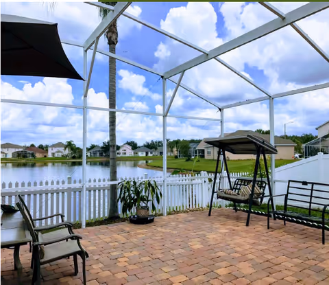 A screened-in patio area with brick flooring, outdoor chairs, a swing bench, and a potted plant. The patio overlooks a small lake with houses and palm trees in the background under a partly cloudy sky.
