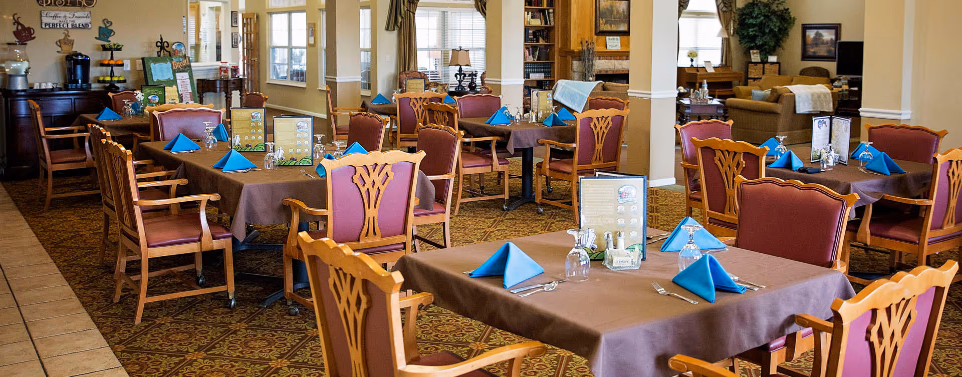 A dining room with multiple tables covered in brown tablecloths and set with blue folded napkins, glasses, and silverware. The room has wooden chairs with red cushions, patterned carpet, and large windows letting in natural light. There are decorative plants and framed pictures on the walls.