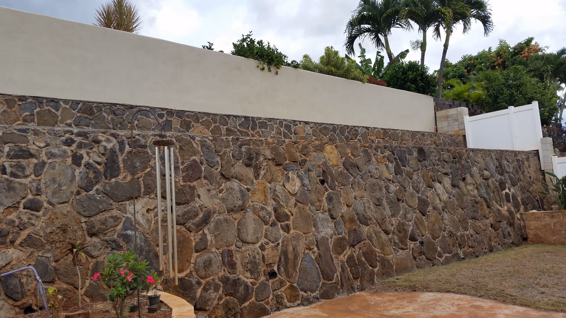 Stone exterior wall topped with a white cap, palm trees, and a dirt yard.