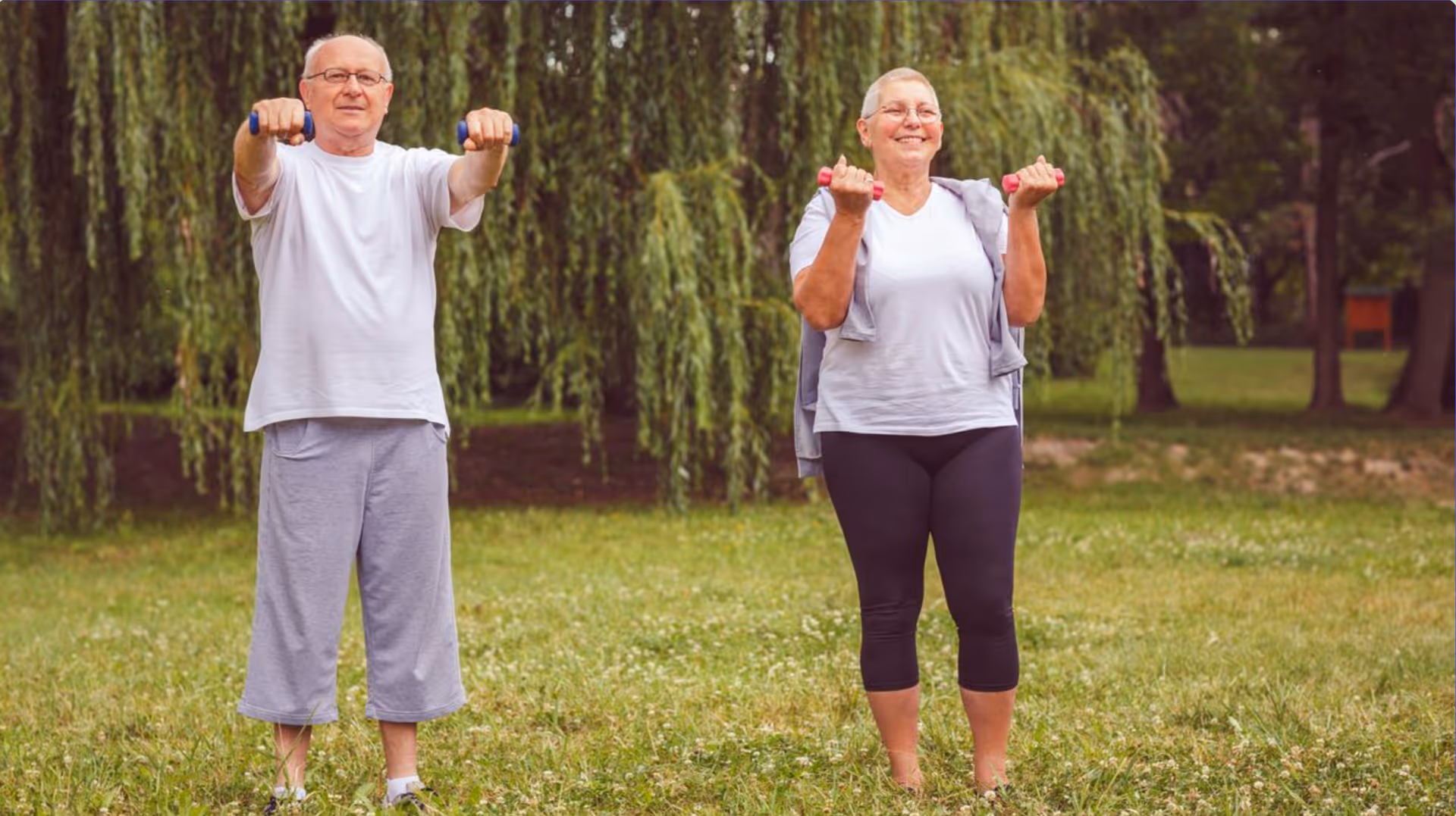 An elderly man and woman exercising outdoors on grass in front of trees, each holding small dumbbells and smiling.