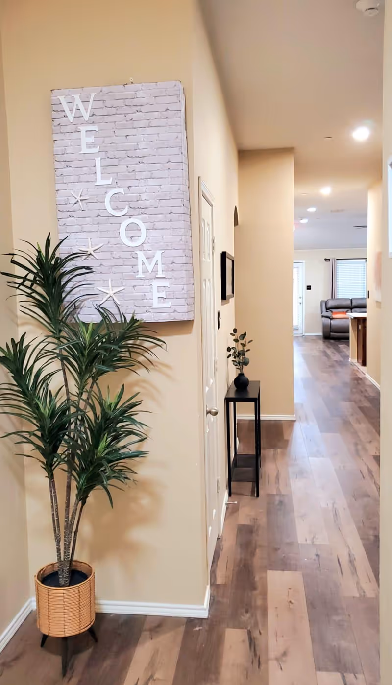 Indoor corridor of Gems Residence with a welcome sign, potted plant, and distant living area.