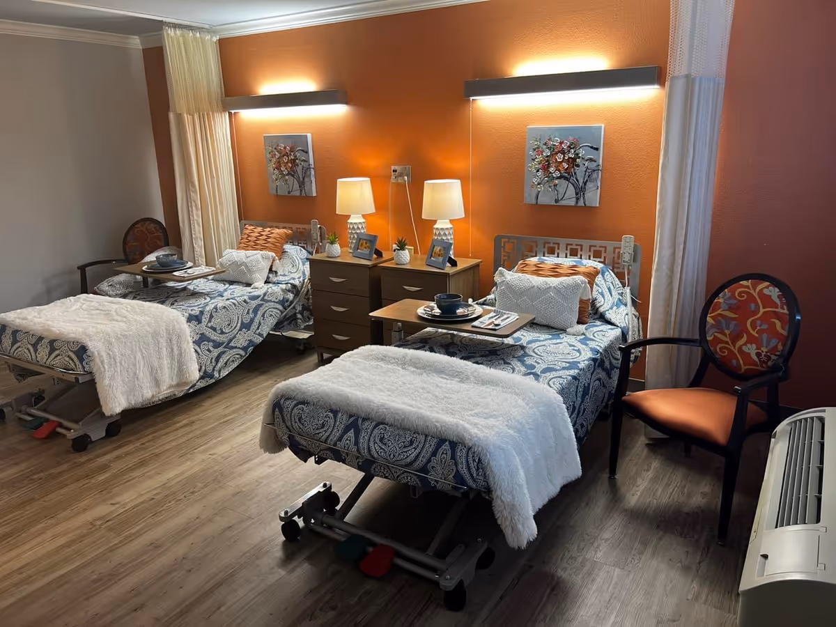 A cozy room with two hospital-style beds, each covered with blue and white patterned bedding and white throw blankets. Each bed has a tray table set with dishes and utensils. Between the beds are two nightstands with table lamps, small plants, and framed photos. The walls are painted orange with floral artwork above each bed. There are two chairs with patterned cushions and a white curtain partially drawn between the beds. The floor is wood laminate.