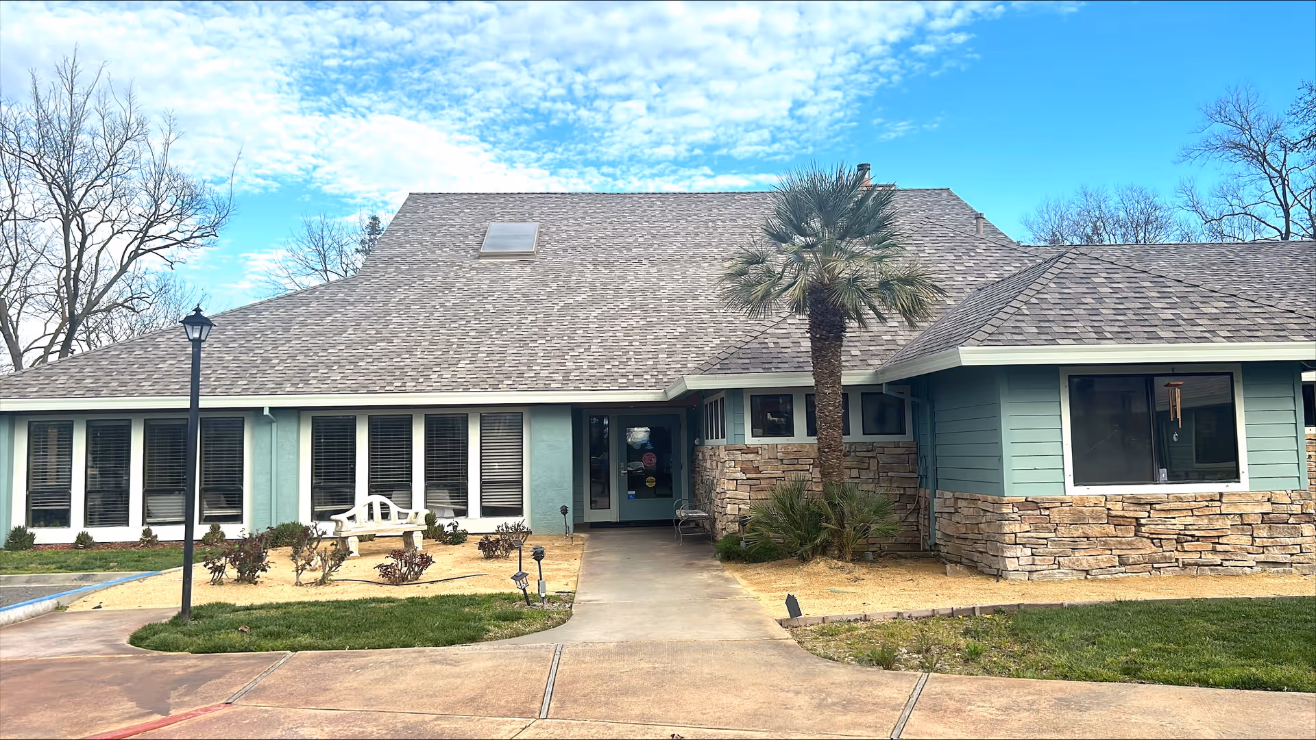 Exterior view of Woodland Gardens Senior Living facility showing a single-story building with a gray shingled roof, light blue walls with stone accents, several windows, a palm tree near the entrance, a white bench, and a clear blue sky with some clouds.