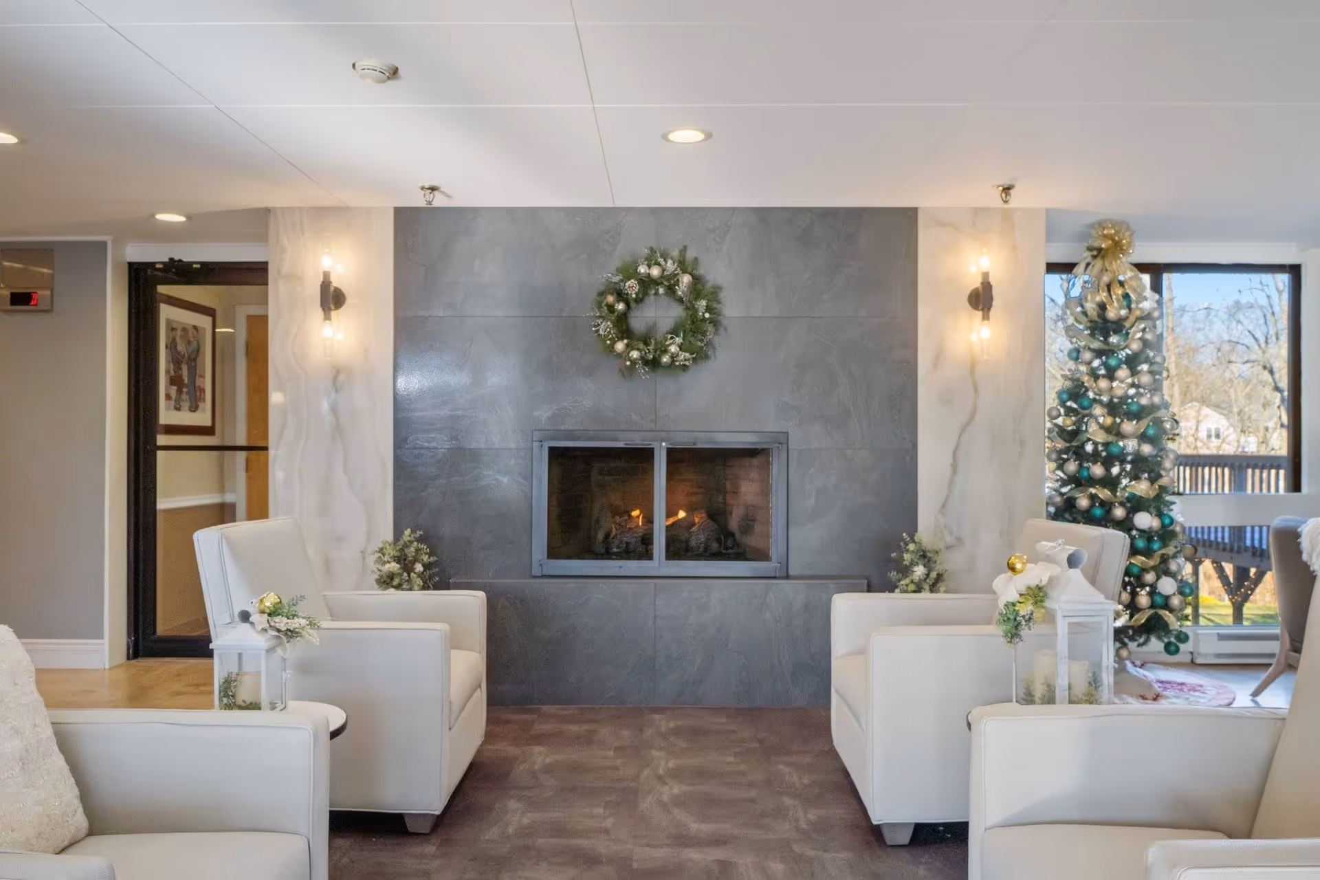 A cozy living room area with a modern fireplace in the center, decorated with a festive wreath above it. There are two white armchairs facing the fireplace, each adorned with small floral arrangements and white lanterns. To the right, a decorated Christmas tree stands near large windows letting in natural light. The room has a warm and inviting atmosphere with soft lighting from wall sconces.