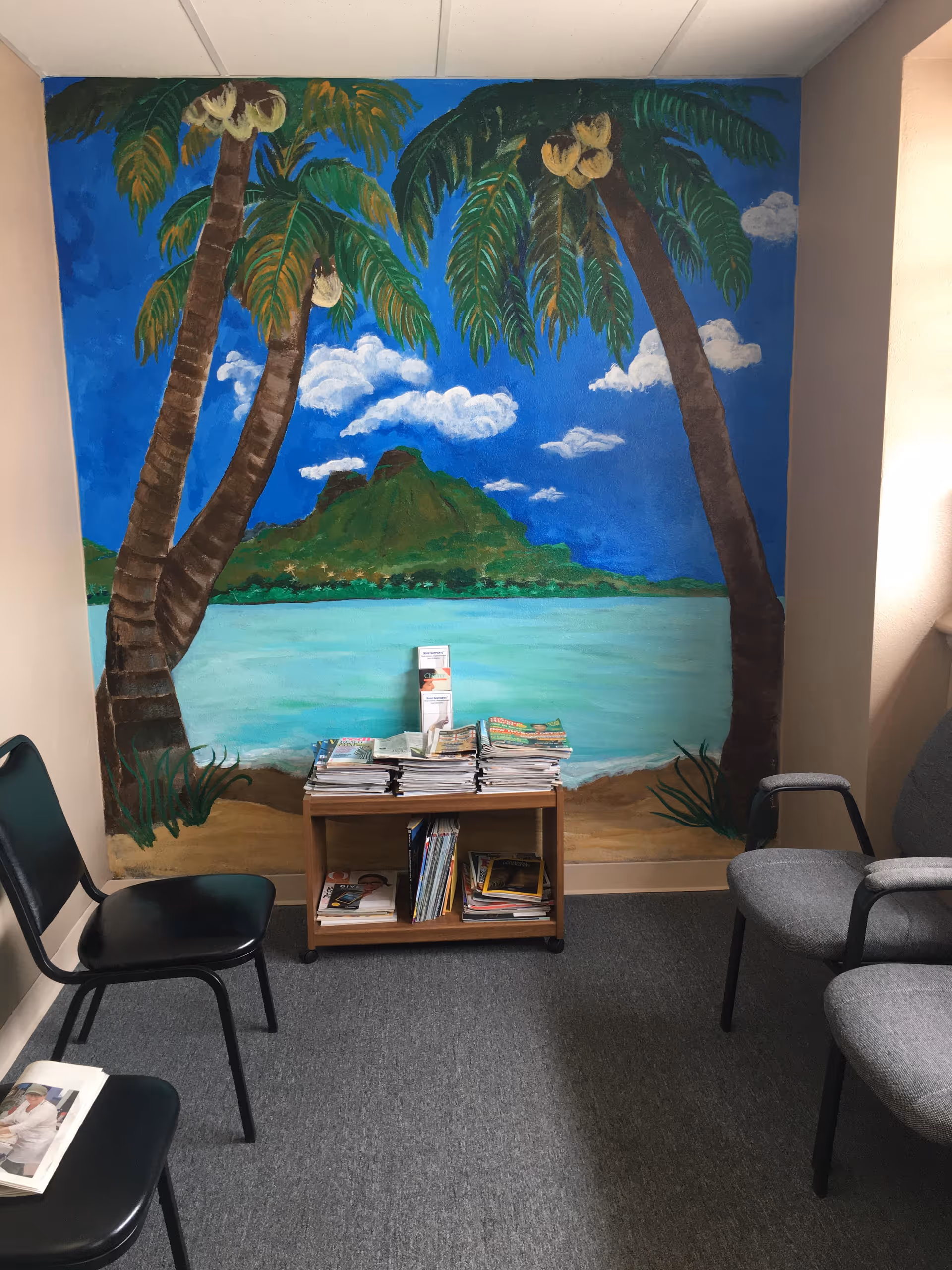 Interior waiting area with a tropical mural, magazine table, and several chairs.