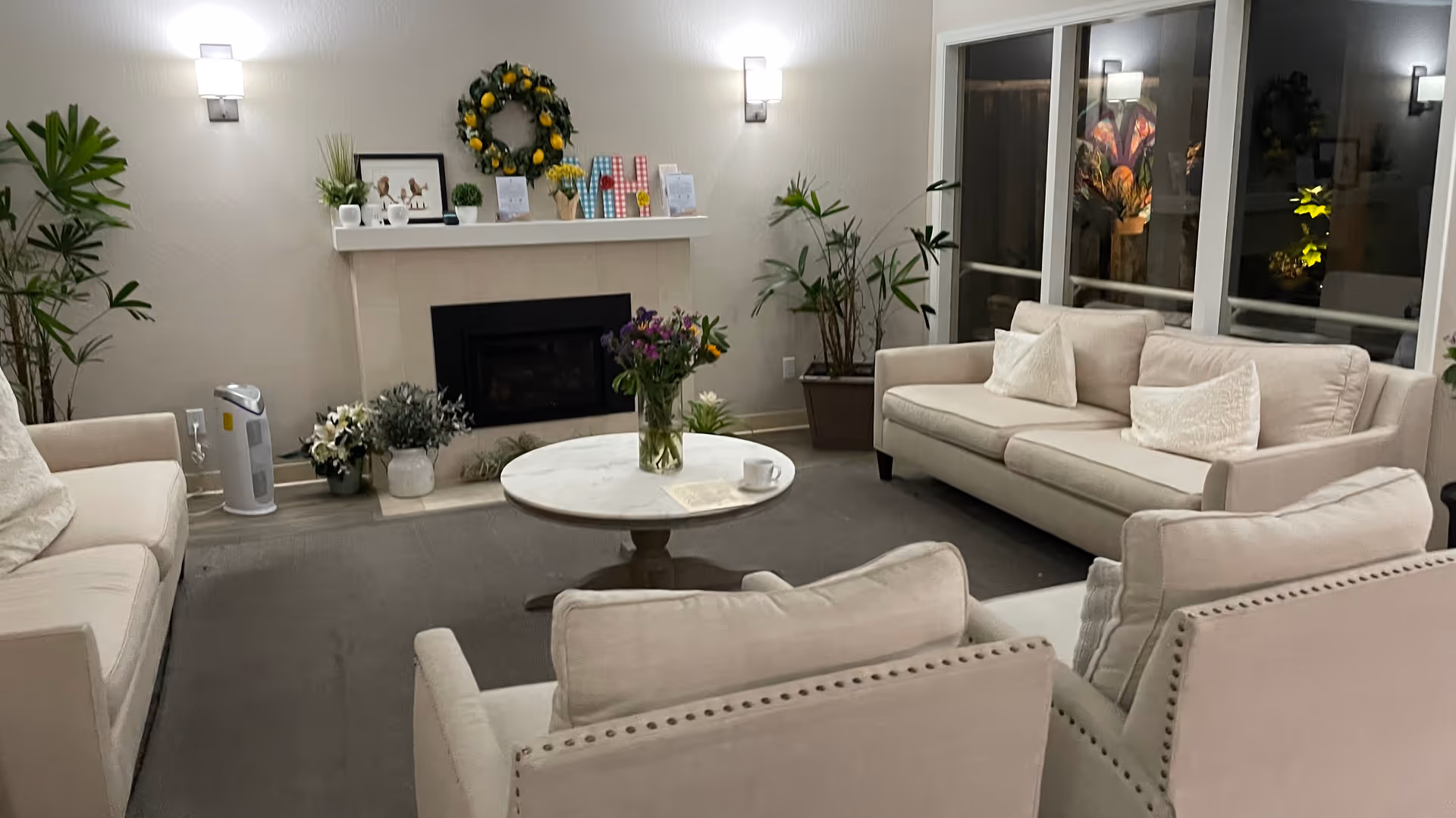 Cozy senior living lounge with beige sofas, plants, a marble coffee table, and a fireplace.