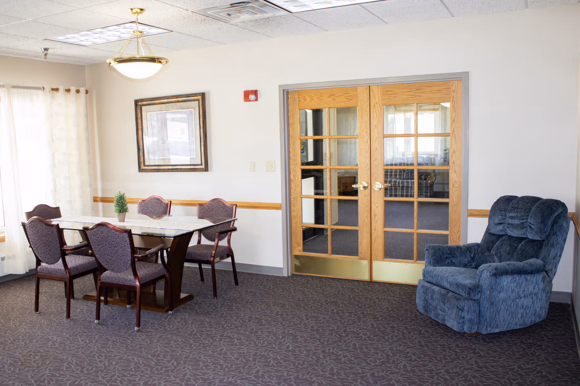 Dining area with glass table, six chairs, blue recliner, and French doors in a senior facility.