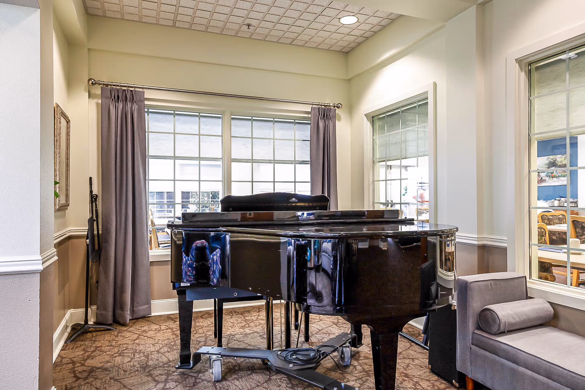 A black grand piano sits in a bright common room with large windows, curtains, and nearby seating.