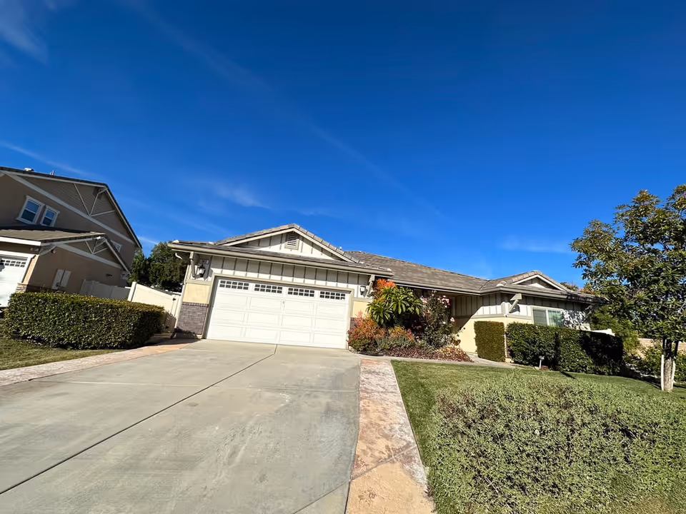 Single-story residential house with a two-car garage, surrounded by neatly trimmed bushes and a green lawn under a clear blue sky.