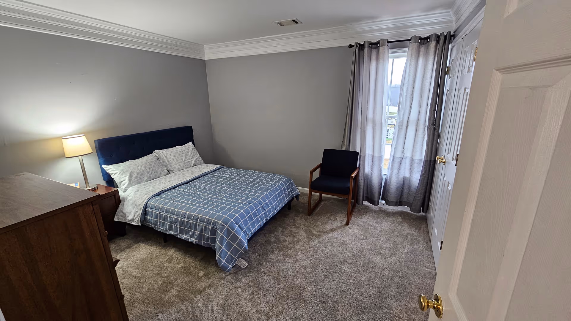 Bedroom with blue-check bed, dresser, lamp, chair, and window with gray curtains.