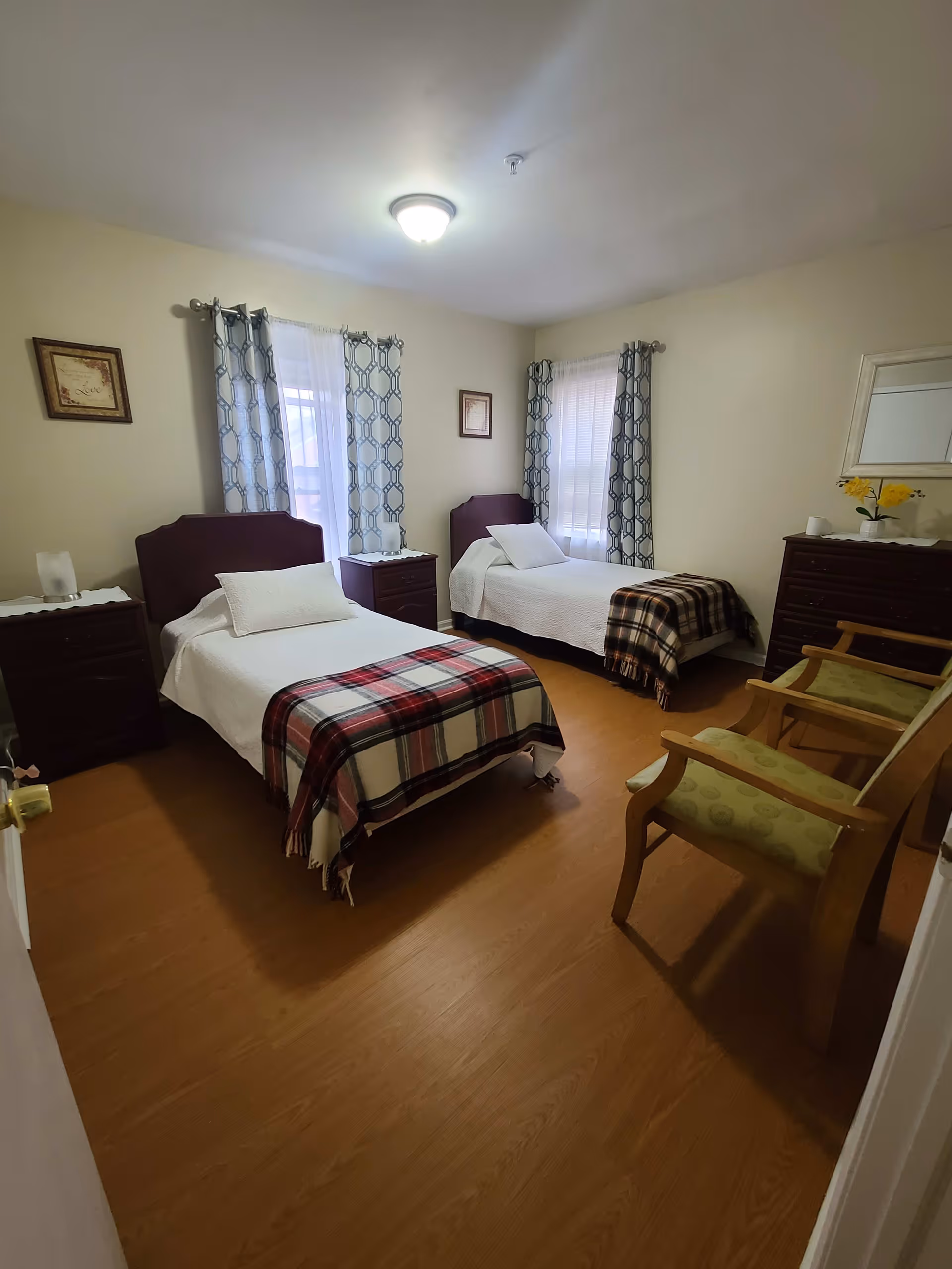 A bedroom with two single beds, each with white bedding and a plaid blanket at the foot. Between the beds is a small nightstand with a lamp. The room has two windows with patterned curtains, light-colored walls, wooden flooring, a dresser with a mirror and yellow flowers, and two wooden chairs with green cushions.