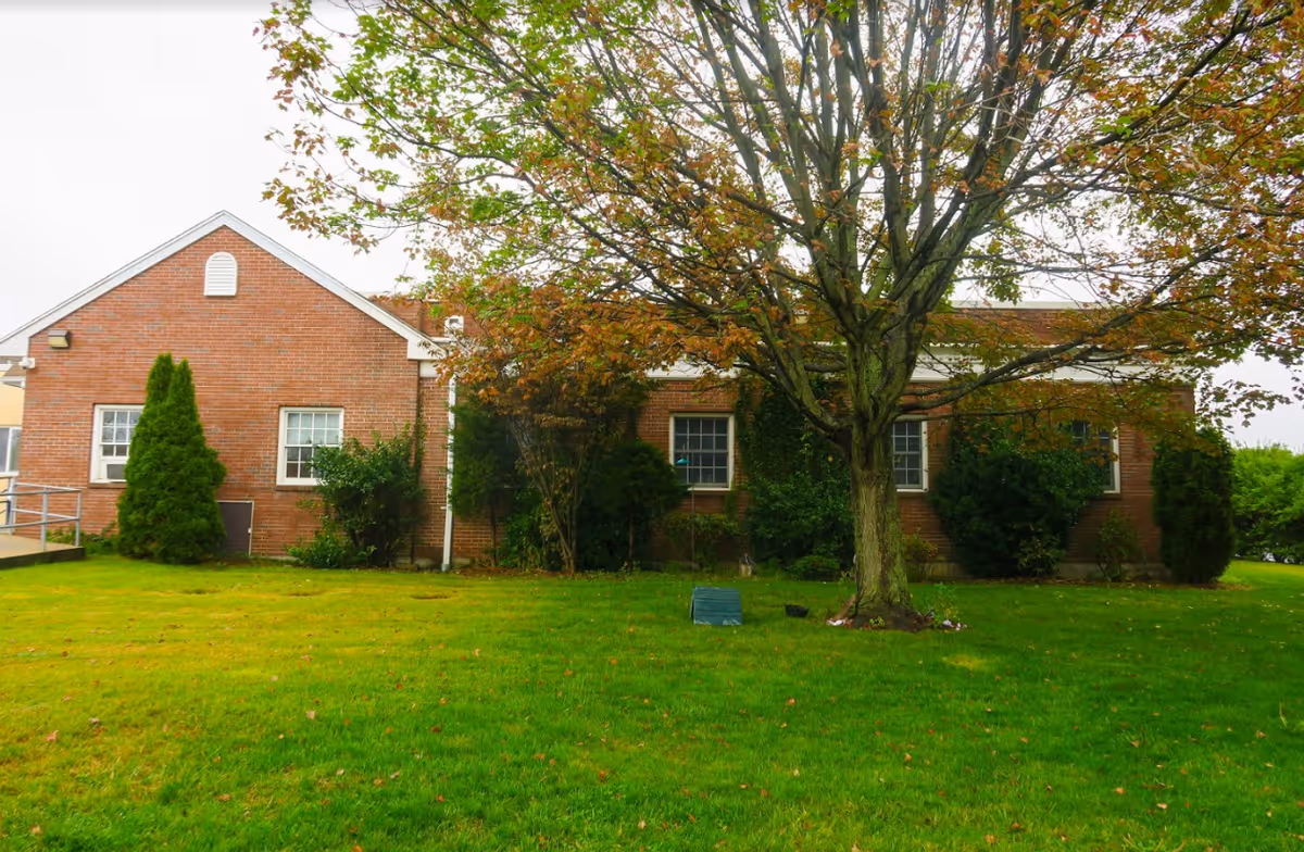 Exterior view of a single-story brick building with several windows, surrounded by green grass, bushes, and a large tree with autumn-colored leaves in the foreground.