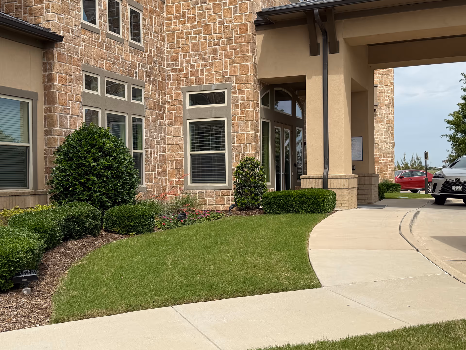 Stone-facade senior living facility entrance with shrubs and parked cars nearby.
