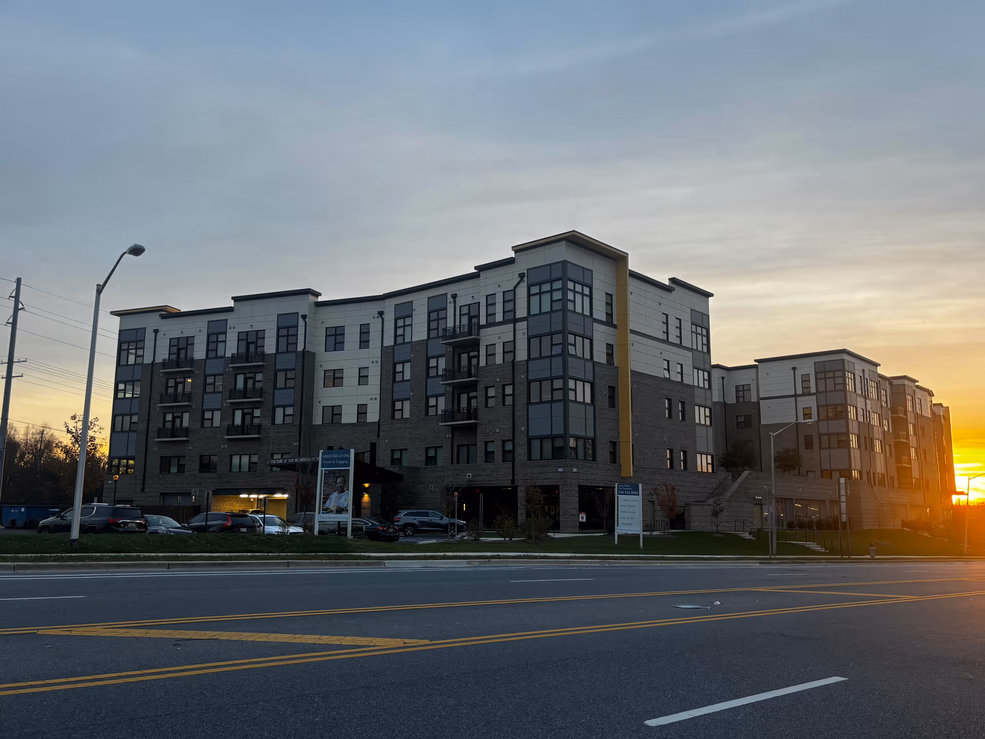Front exterior of a multi-story senior living building at sunset with parked cars along the street.