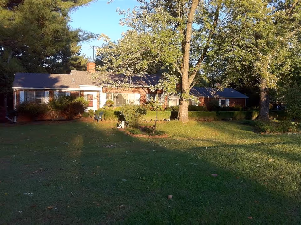 Single-story brick ranch-style building with a large front lawn, trees, and shrubs in sunlight.