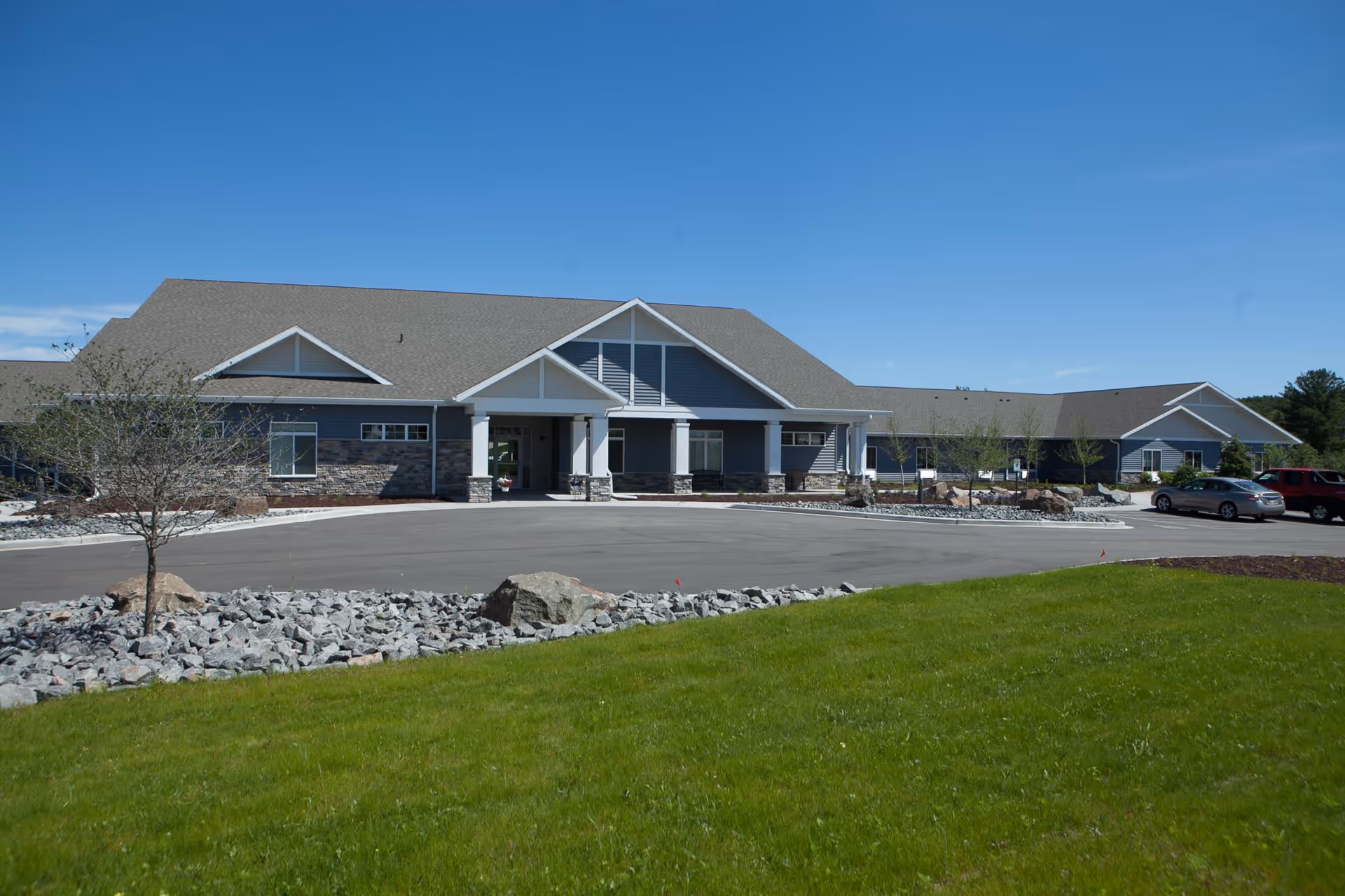 Front exterior view of a single-story assisted living facility building with a large covered entrance, stone and blue siding, surrounded by a paved driveway, landscaped rocks, small trees, and green grass under a clear blue sky.
