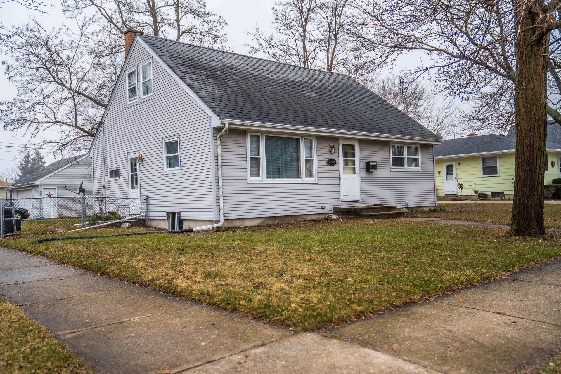 Single-story light gray house with a sloped roof, front door, and a small lawn on a residential corner lot.