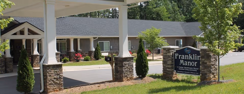 Exterior view of Franklin Manor Assisted Living Center showing the entrance with white columns and stone bases, a landscaped area with small trees and bushes, and a sign displaying the facility's name and description.