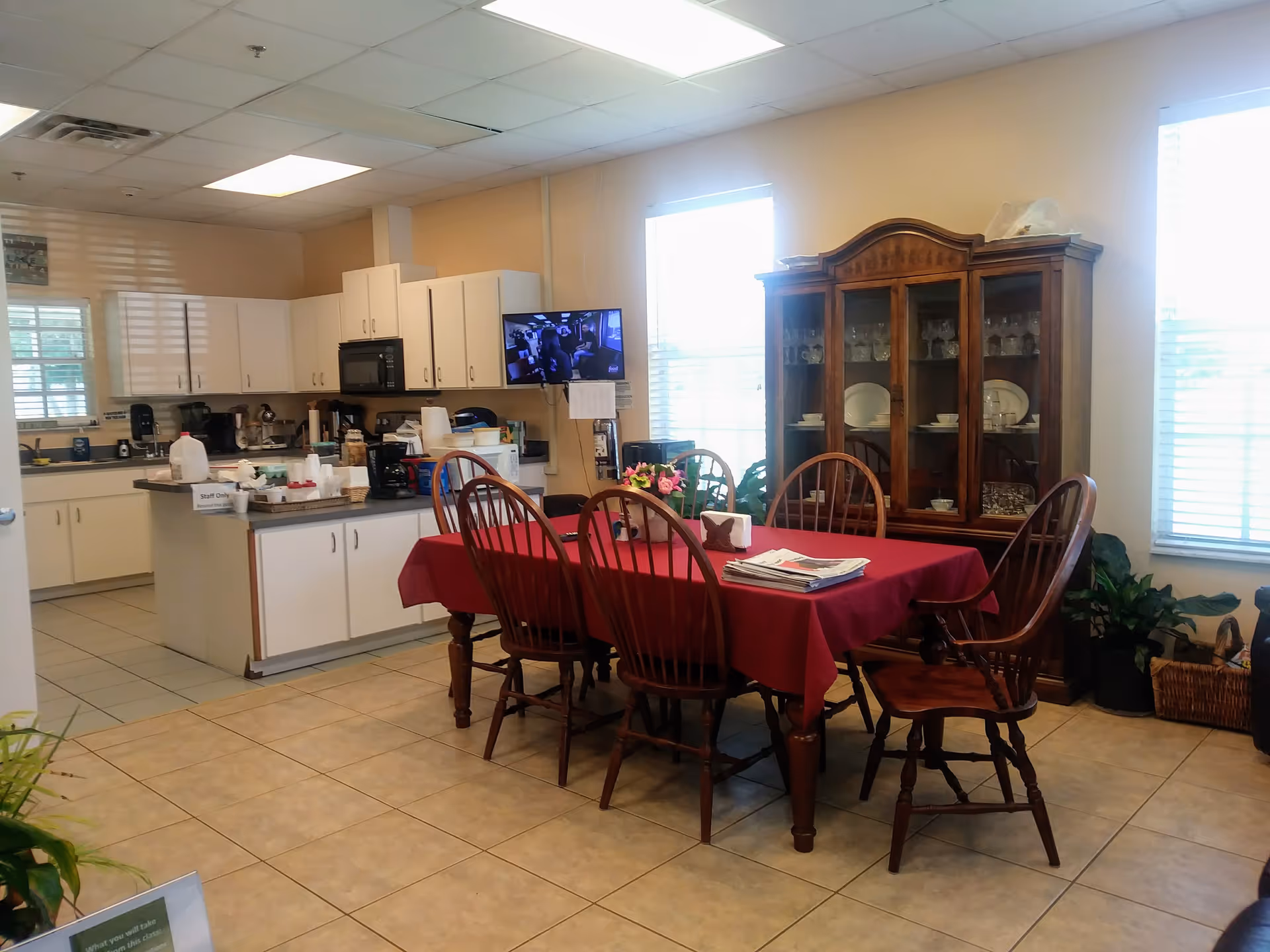 Dining room with a red tablecloth, wooden chairs, and a china cabinet beside a kitchen.