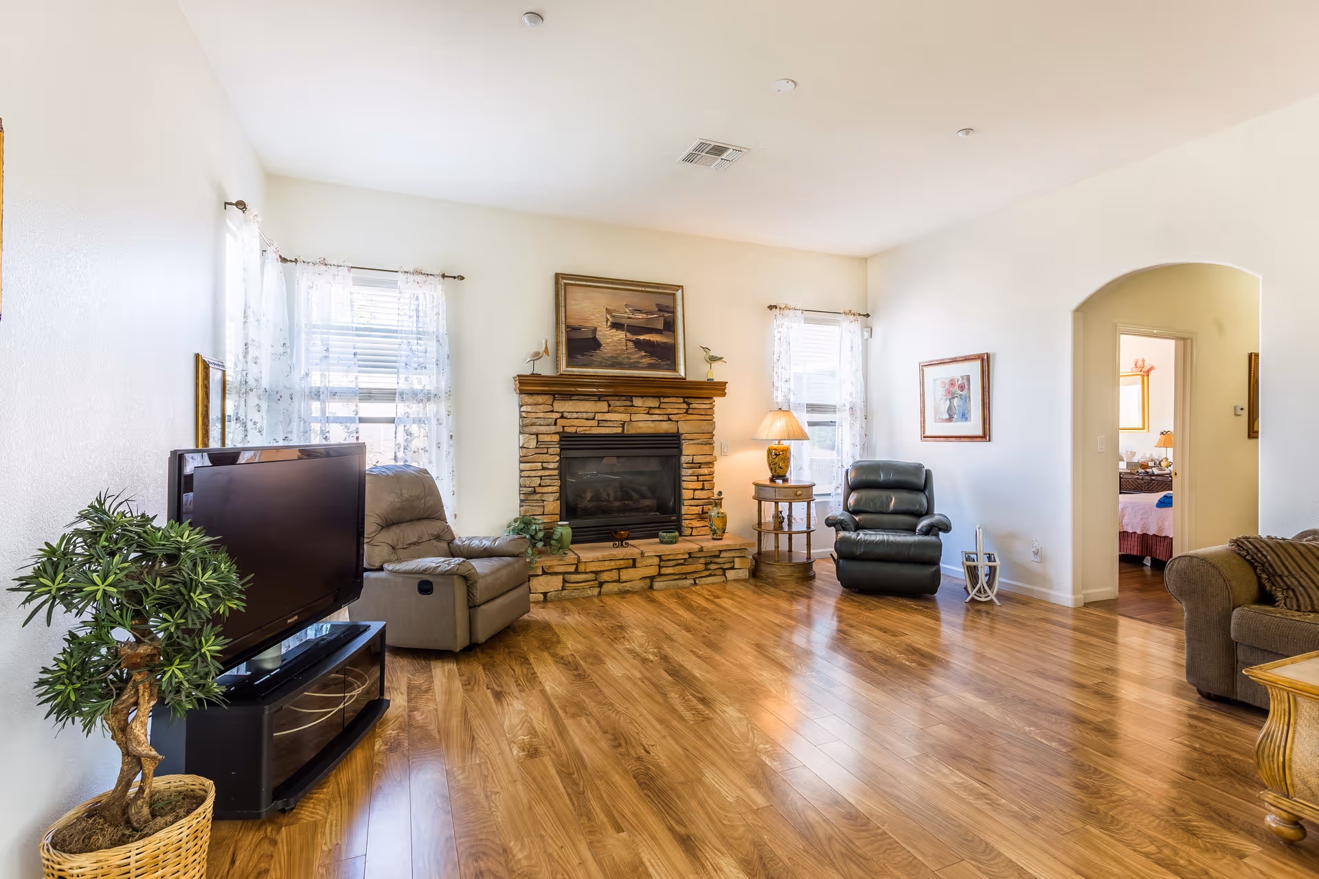 A bright living room with wooden flooring, a stone fireplace with a painting above it, two armchairs, a TV on a stand, a potted plant, a side table with a lamp, and two windows with sheer curtains. An open doorway leads to another room with a bed visible.