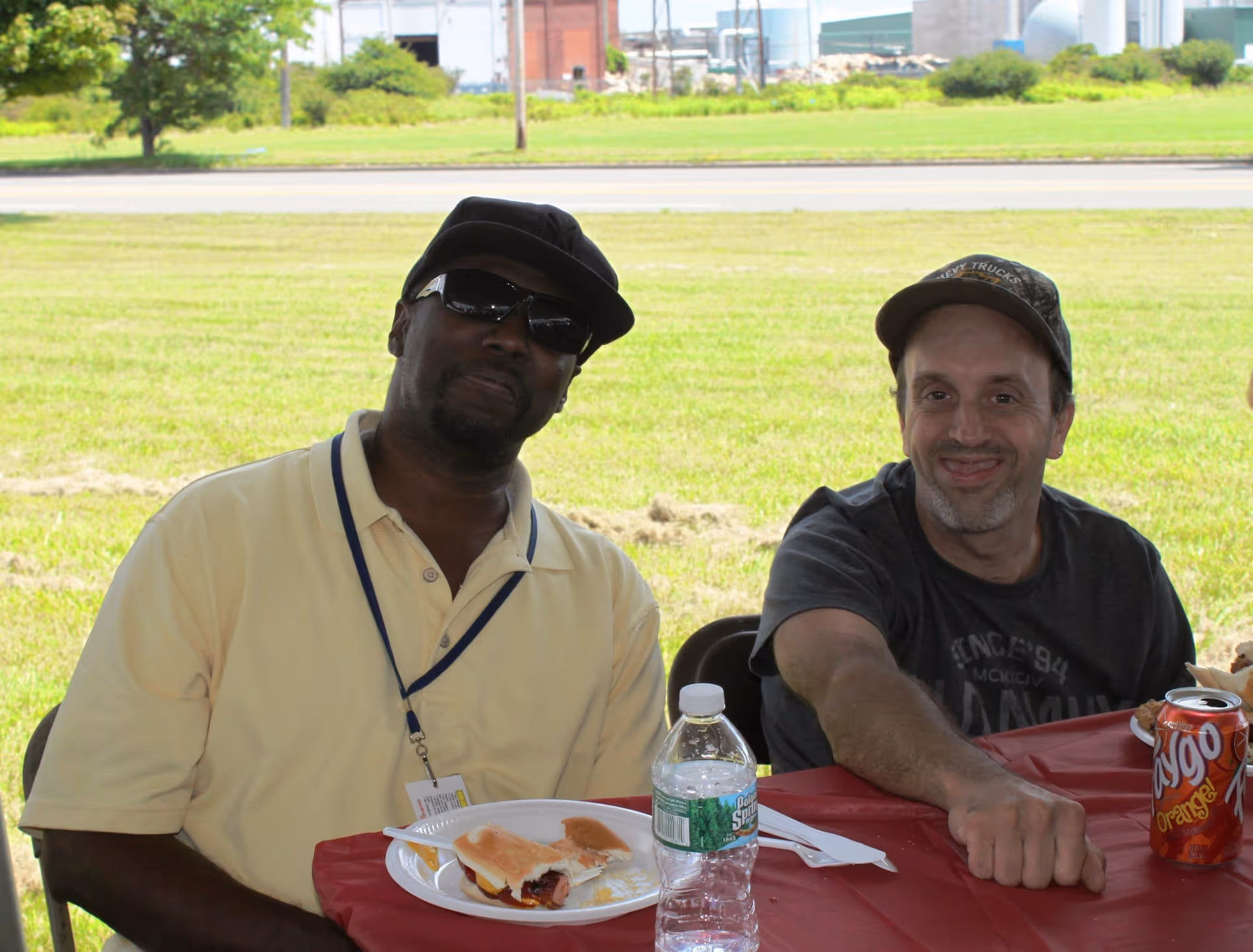 Two men sit at a red outdoor table, eating sandwiches and drinking beverages.