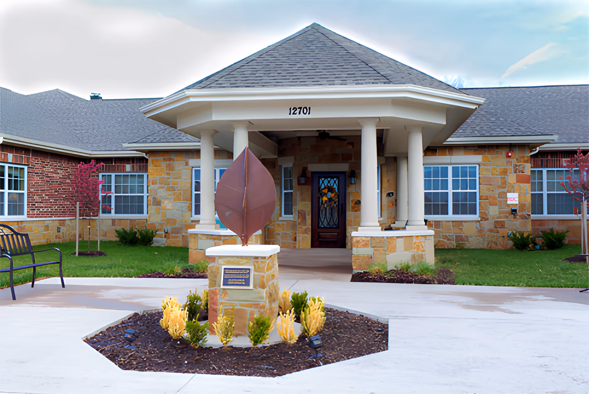 Front entrance of a single-story brick and stone memory care building with a covered portico supported by columns, a decorative leaf sculpture on a pedestal in the foreground, and landscaped grounds.