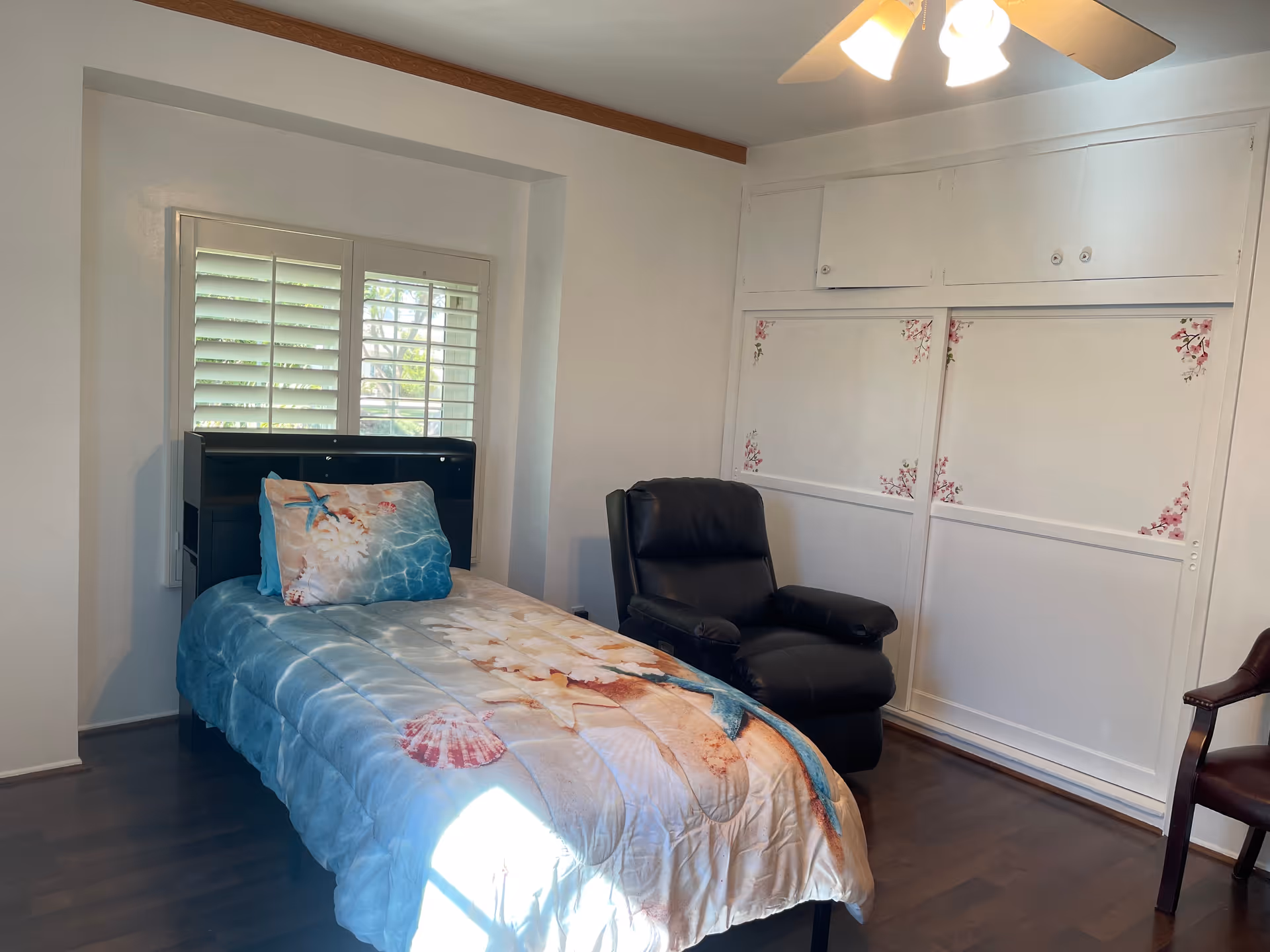 A small bedroom with a single bed, recliner chair, window with shutters, and built-in white cabinets.