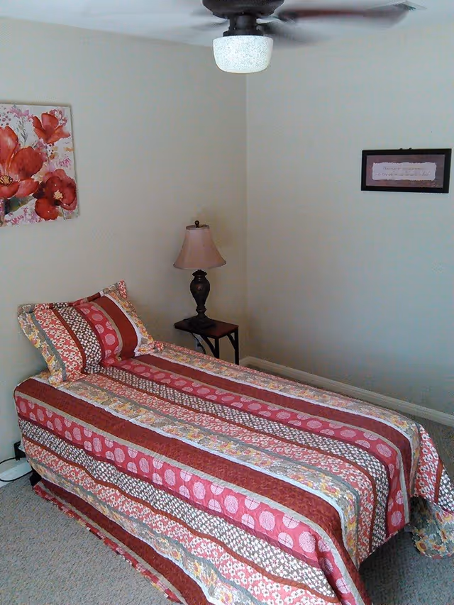 A small bedroom with a pink patterned quilt on a single bed, lamp, and wall art.