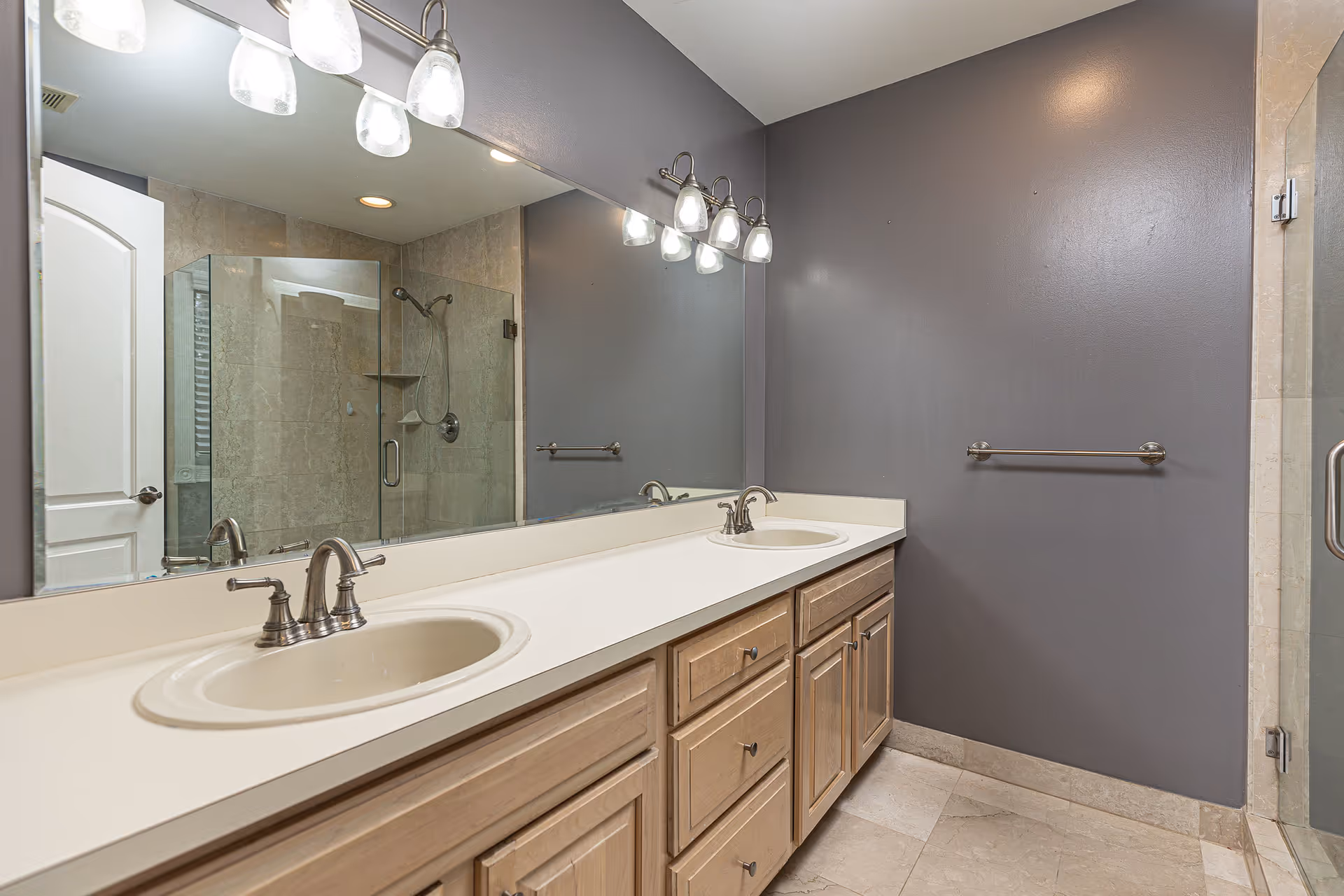 Double-sink bathroom vanity with large mirror, glass shower, and neutral tiled floor.